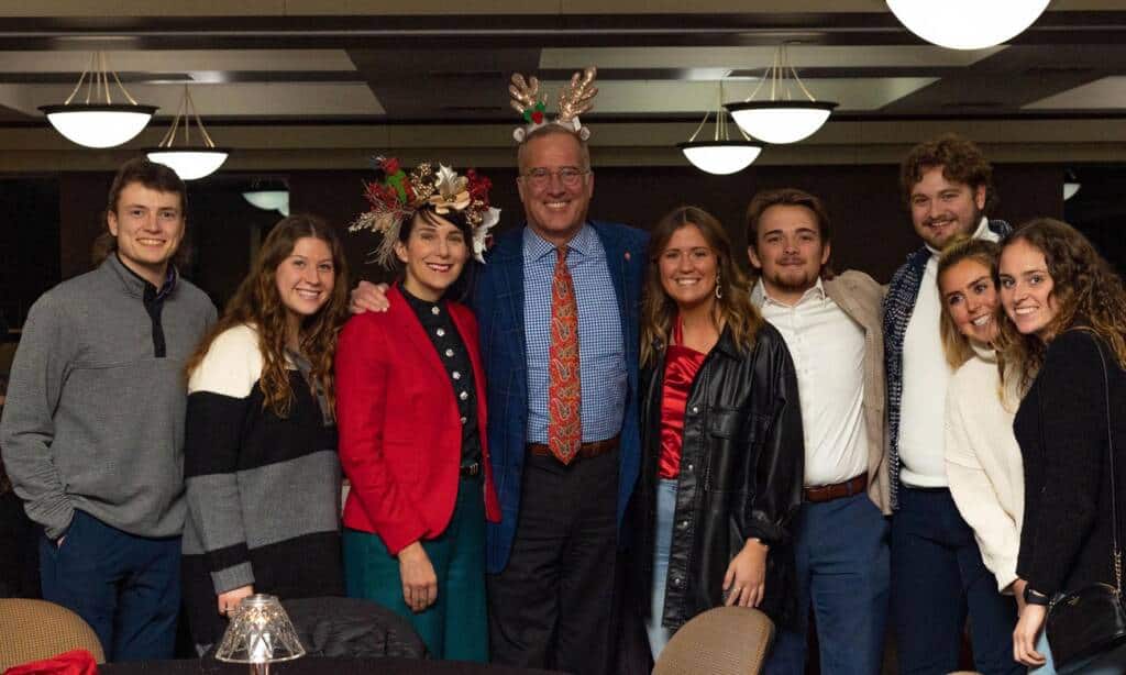 A group of nine people smiling and posing for a photo indoors. Two individuals in the center are wearing festive reindeer headbands. They stand closely together under ceiling lights in a warmly lit room, suggesting a celebratory or social event.