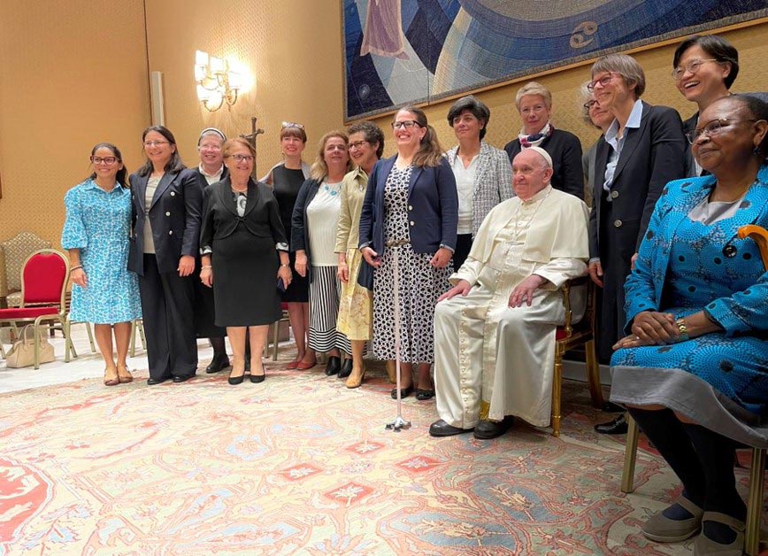 A group of women, dressed in formal attire, stands in a semicircle around a seated Pope in a white robe. They are indoors, in a room with an ornate carpet and a large decorative wall hanging in the background. All are smiling for the photo.