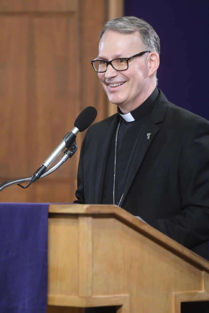 A man wearing glasses and clerical attire stands behind a wooden podium, smiling while speaking into a microphone. The background includes wooden paneling and a dark blue fabric.