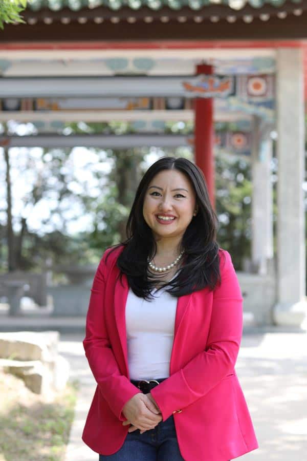 A woman with long black hair, wearing a pink blazer over a white shirt and blue jeans, stands smiling in an outdoor setting with traditional East Asian architecture in the background. Trees and sunlight are also visible.