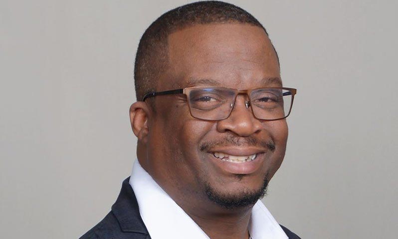 A man with short hair and glasses is smiling at the camera. He is wearing a dark blazer over a white collared shirt, and the background is plain and light-colored.