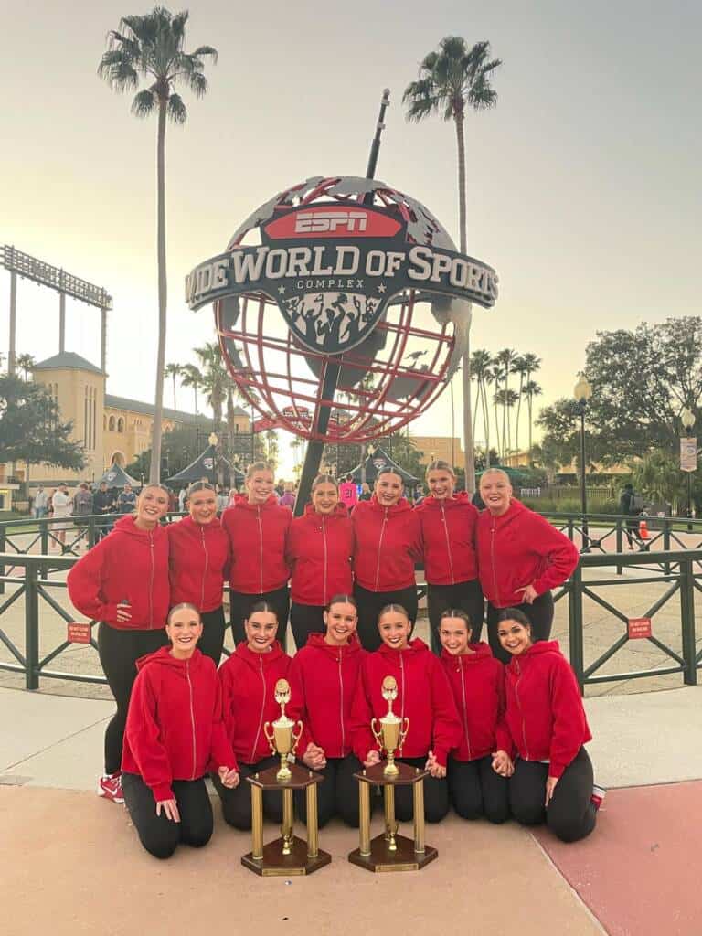 A group of cheerleaders in matching red jackets and black pants pose with three trophies in front of the ESPN Wide World of Sports Complex sign. Palm trees and a clear sky are in the background.