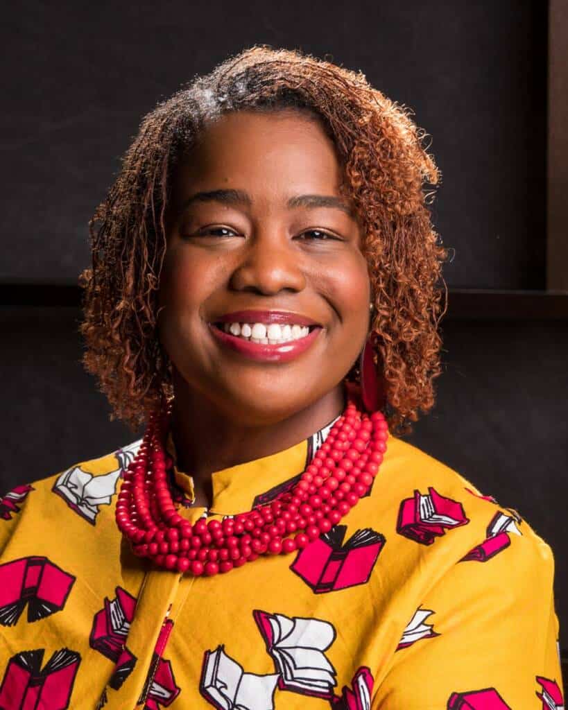 A woman with curly hair smiles warmly at the camera. She is wearing a brightly colored yellow blouse with red and white book patterns, a vibrant red beaded necklace, and matching earrings. The background is dark, making her colorful outfit stand out.