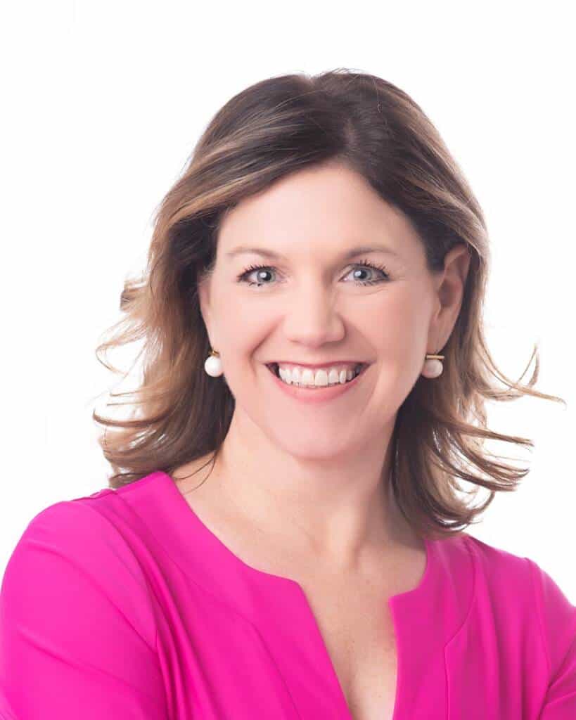 A smiling woman with shoulder-length brown hair, wearing a bright pink top and pearl earrings, poses against a plain white background.