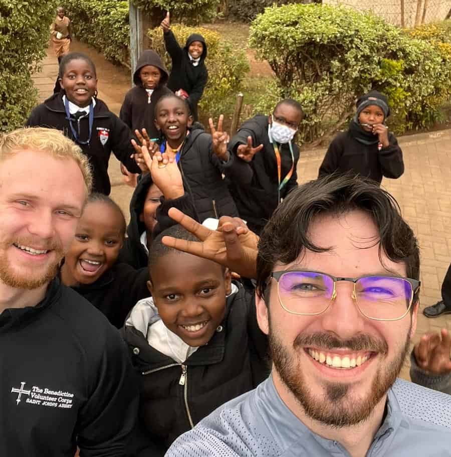 A group of children and two adults smile at the camera outdoors. The children are wearing black clothing and making peace signs. One of the adults wears glasses and a grey jacket, and the other has a beard and a black jacket with text on it. Trees and bushes are in the background.