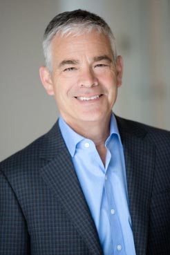 A middle-aged man with short, gray hair is smiling. He is wearing a blue dress shirt and a dark checked blazer. The background is neutral and slightly blurred.