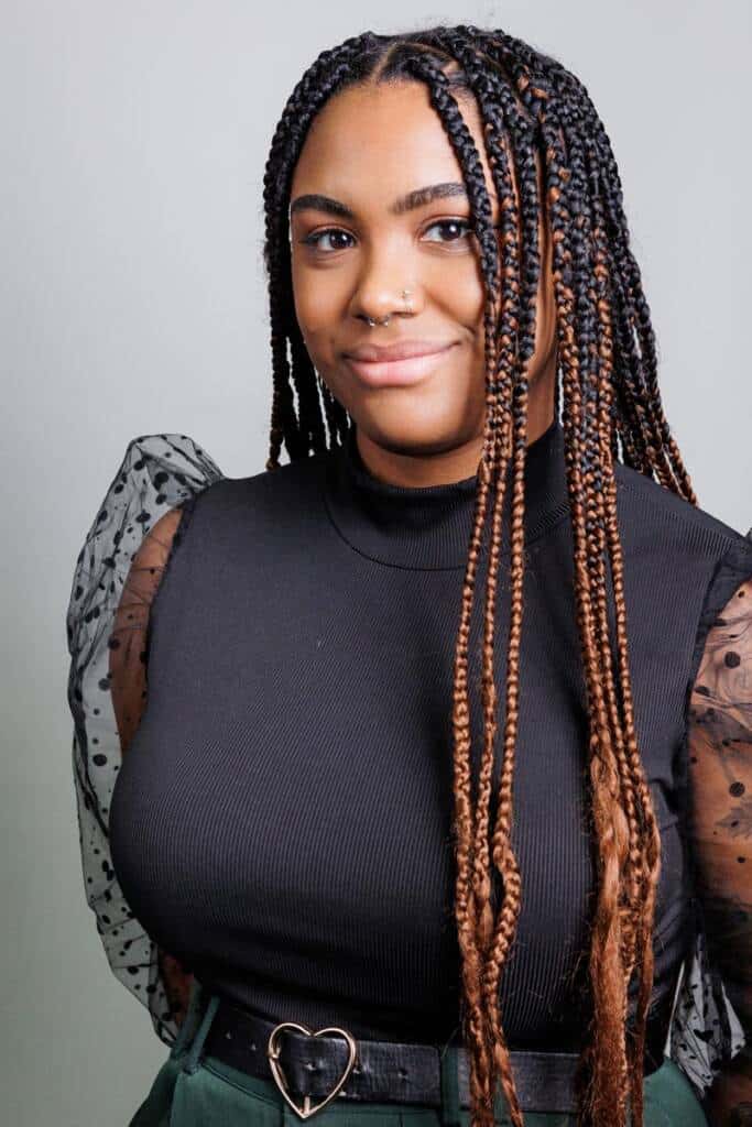 A woman with long braided hair smiles softly at the camera. She is wearing a black top with polka dot mesh sleeves and a high neck, paired with green pants featuring a belt with a heart-shaped buckle. The background is a plain light gray color.