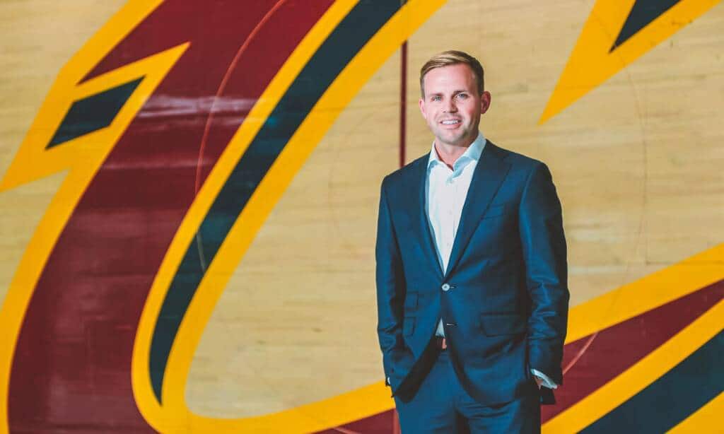 A man in a dark suit stands confidently in front of a large Cleveland Cavaliers logo on a wooden floor. He has short hair and is smiling, with his hands in his pockets. The background features the team's distinctive maroon and gold colors.