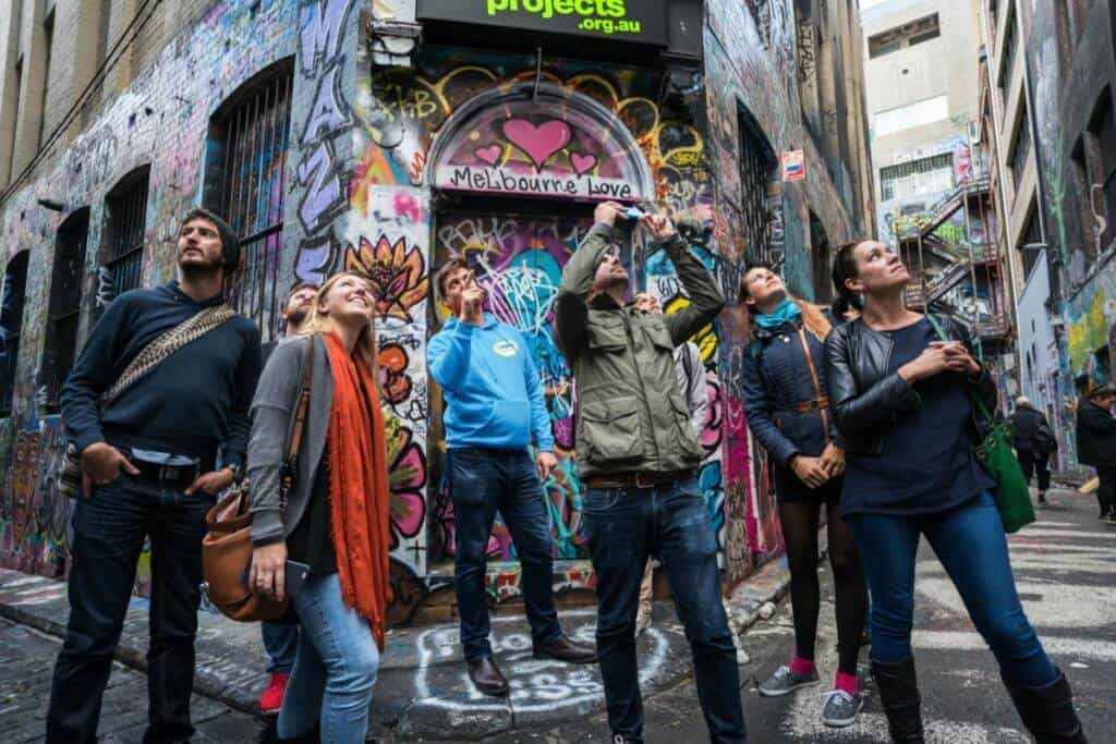 A group of people stands in a colorful graffiti-covered alley, looking up at the walls. They appear to be on a guided tour. The alley includes various vibrant street art, with a sign above reading "Melbourne Love." The mood is lively and curious.