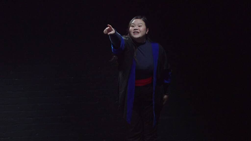 A person dressed in a dark robe with blue accents and a red sash performs on stage. They are standing against a dark background, extending their right arm and pointing forward, with an expressive facial expression.