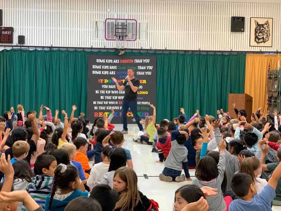 A person stands on a stage in front of a large group of children seated on a gym floor. The children have their hands raised. A motivational banner with colorful text hangs behind the person. The background includes gym equipment and a scoreboard.