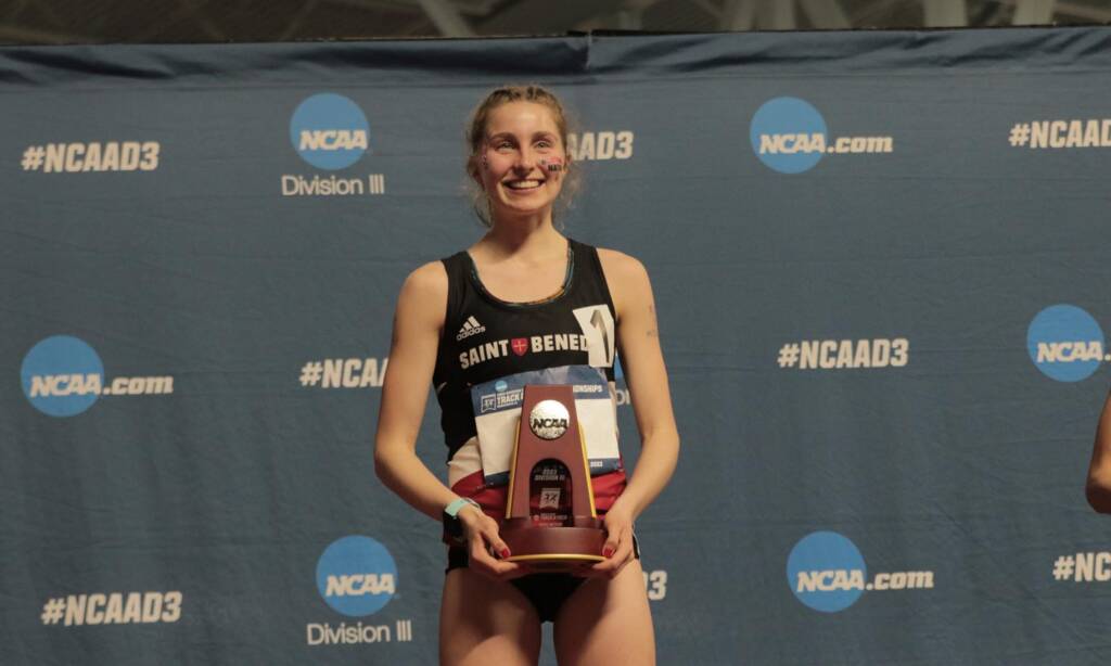 A female athlete wearing a "Saint Benedict" jersey stands smiling while holding a trophy in front of a backdrop with NCAA logos and "#NCAAD3" text.