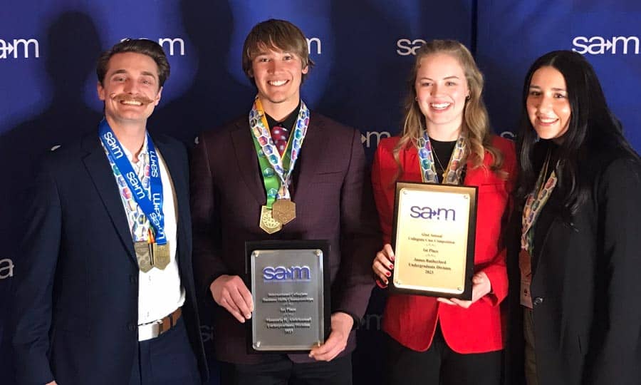 Four smiling individuals stand in front of a blue backdrop with the word "samm" repeated on it. Two of them hold plaques, and two of them have medals around their necks. They appear to be at an award event, and everyone is dressed in formal attire.