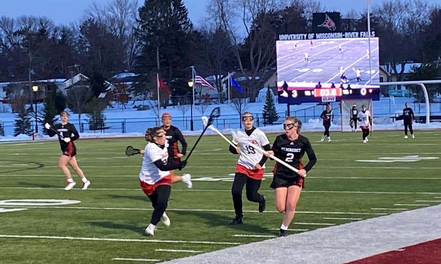 A women's lacrosse game is underway on a field with snow in the background. One team wears white jerseys and the other black, with players in action. A scoreboard in the distance shows University of Wisconsin - River Falls, displaying a close game score.