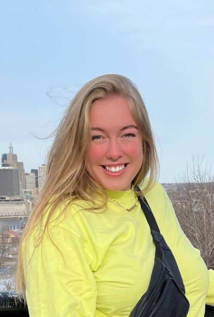 A person with long, blonde hair smiles brightly at the camera. They are wearing a yellow top and a black sling bag. The background features a cityscape with skyscrapers and a clear, blue sky.