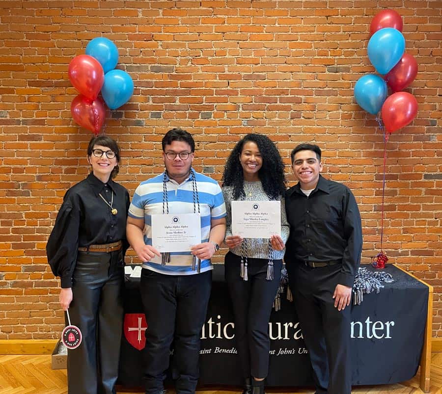 Four people stand together in front of a brick wall and a table with the word "Center" on it. Two people in the middle hold certificates. Red and blue balloons are tied to the table’s corners. The group is smiling.