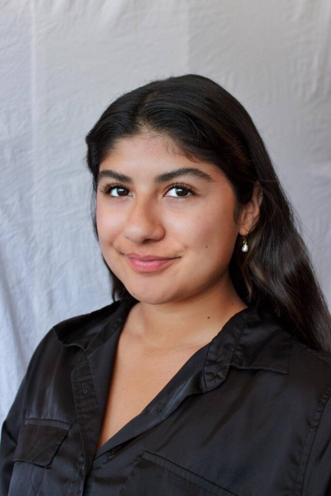 A woman with long dark hair, wearing a black shirt and small hoop earrings, is smiling softly at the camera. The background is plain white fabric.