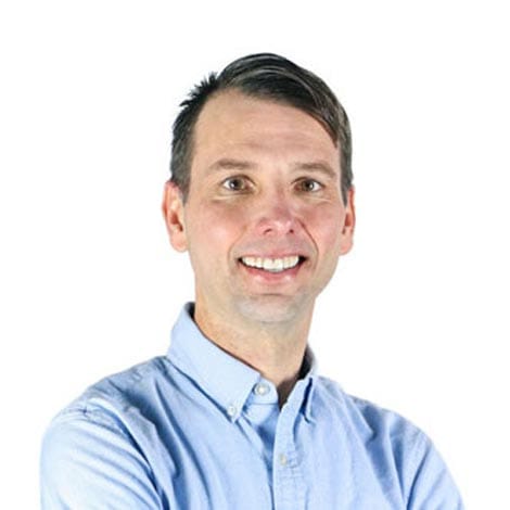 A man with short dark hair and light skin is smiling, looking directly at the camera. He is wearing a light blue button-down shirt and is standing against a plain white background. His arms are crossed casually in front of him.