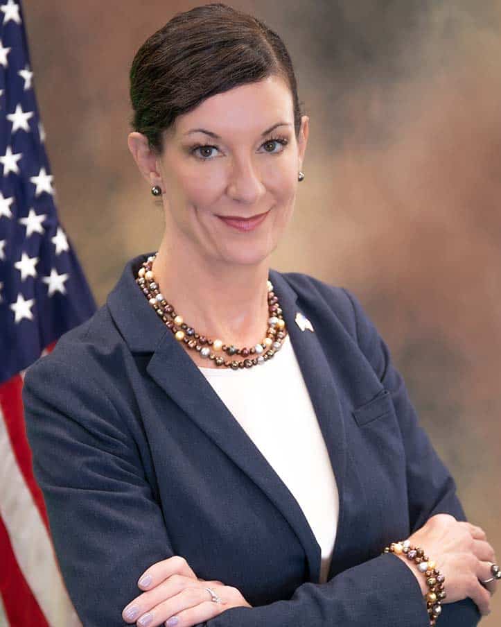 A woman with short dark hair stands with her arms crossed, wearing a navy blazer, white blouse, and beaded necklace and bracelet. She is smiling and standing in front of an American flag and a neutral blurred background.