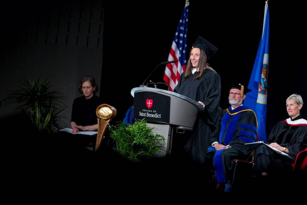 A person in graduation attire speaks at a podium on stage with the College of Saint Benedict emblem. Seated behind are other officials in academic robes. American flag and another flag are in the background. A potted plant and ceremonial mace are on stage.