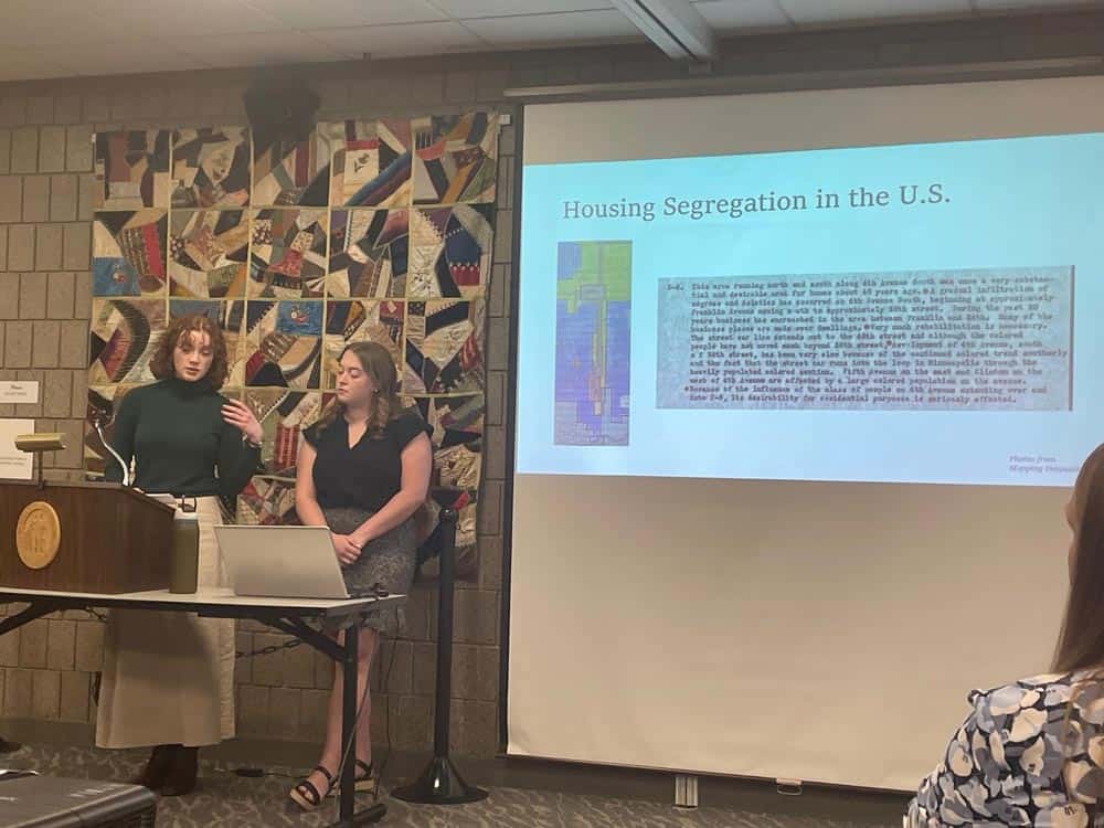 Two individuals are giving a presentation in a room with a patchwork quilt on the wall. They are discussing "Housing Segregation in the U.S.," as displayed on a projection screen featuring an old document and a colored map. One person speaks at a podium, while the other sits beside a laptop.