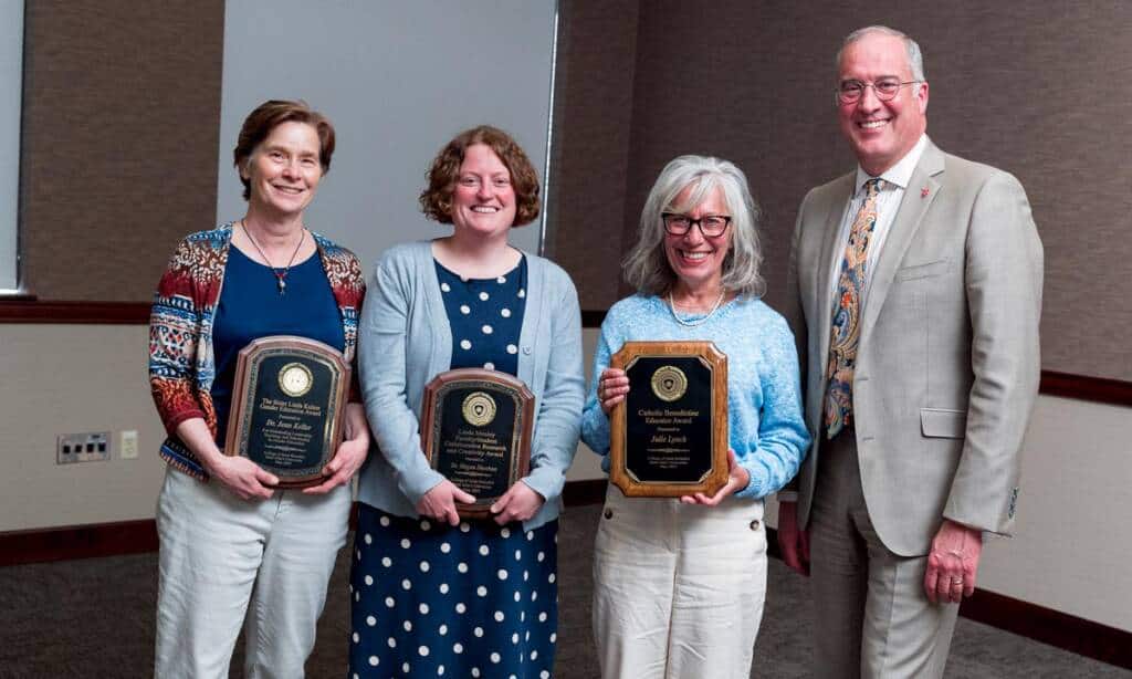 Four people are standing indoors, smiling at the camera. Three people are holding plaques with black and gold detailing. They are dressed in business casual attire. The backdrop includes a beige wall and a gray projection screen.