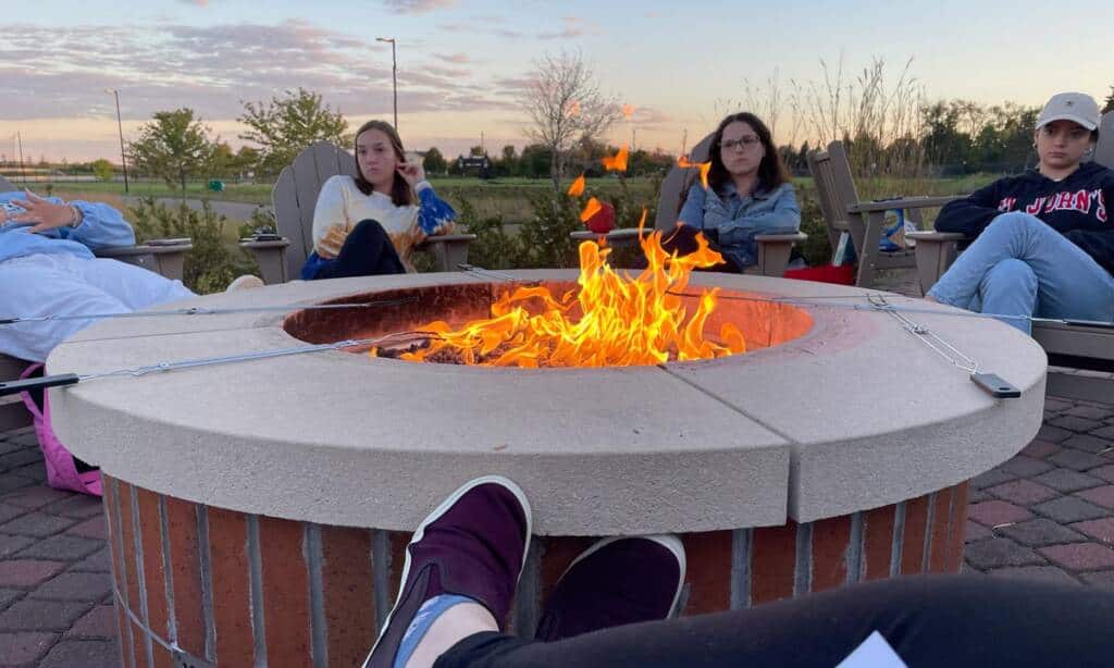 Four people seated around a circular, brick fire pit with a bright, blazing fire in the center. They are sitting on wooden chairs outdoors, with one person resting their feet near the edge of the pit. The sky is partly cloudy and there are surrounding trees and shrubs.