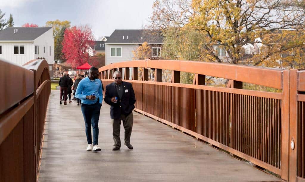 Two men walk side by side on a bridge, engaged in conversation. The bridge is brown with railings, and autumn trees with colorful leaves surround the area. In the background, other people can be seen walking, with residential buildings lining the scene.