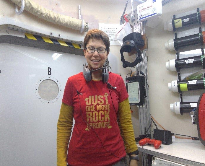 A person wearing glasses and a red t-shirt with the text "JUST ONE MORE ROCK I PROMISE" stands in a room with various technical equipment. They have headphones around their neck and appear to be in a professional or lab setting.