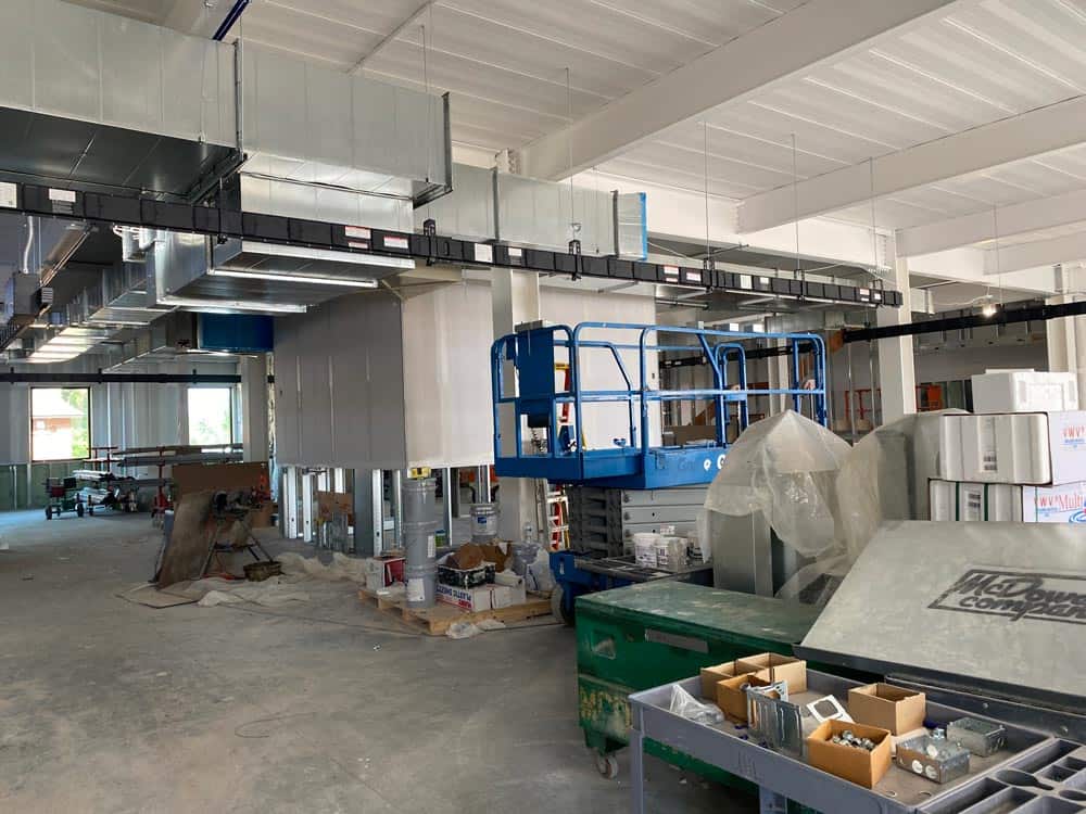 A large industrial space under construction with exposed ducts, partially installed partitions, and construction materials scattered around. A blue scissor lift is parked in the center, surrounded by tools, boxes, and pieces of insulation.