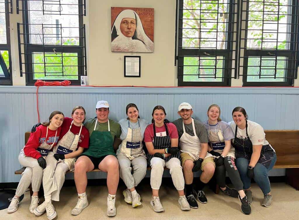 A group of eight people dressed casually in aprons sit on a bench indoors, smiling at the camera. Behind them are large windows, and a painting of a nun is mounted on the wall. They appear to be in a light-filled, communal space.