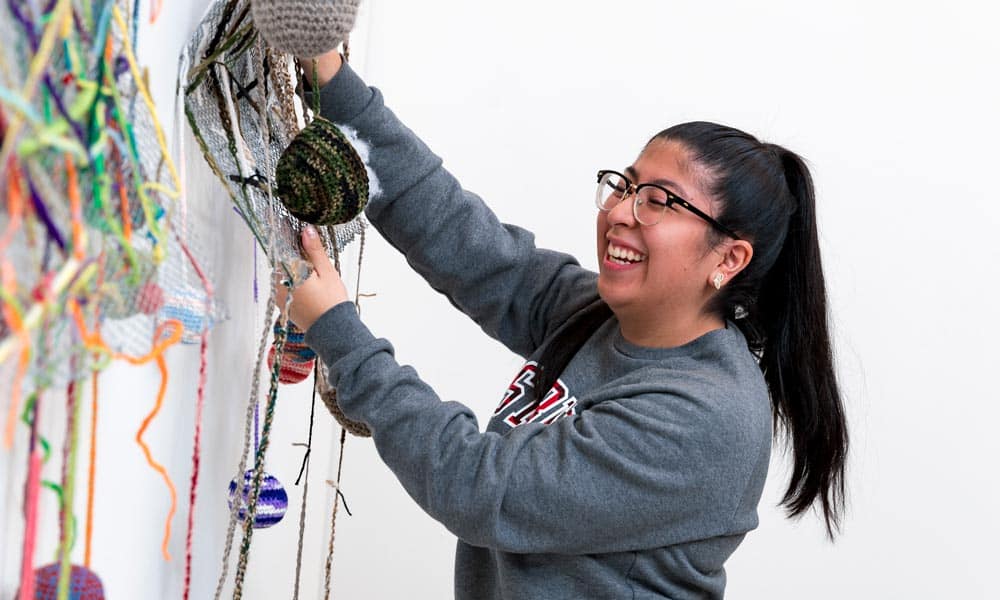 A woman with glasses, wearing a gray sweatshirt, is smiling as she arranges colorful yarn art on a wall. She has long dark hair tied back in a ponytail and is engaging with the creative, vibrant display.