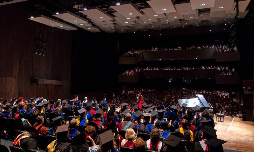 A large auditorium filled with people attending a graduation ceremony. Graduates in caps and gowns are seated on the stage while faculty members, also in academic regalia, are seated behind them. The audience occupies the seats on multiple levels of the auditorium.