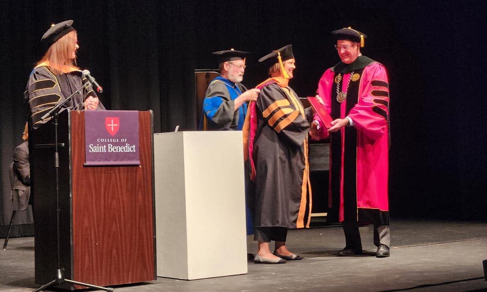 A graduate in black and gold academic regalia shakes hands with a faculty member in pink regalia while receiving a diploma on a stage. Another faculty member assists with the hood. A woman at a podium that reads "College of Saint Benedict" looks on.