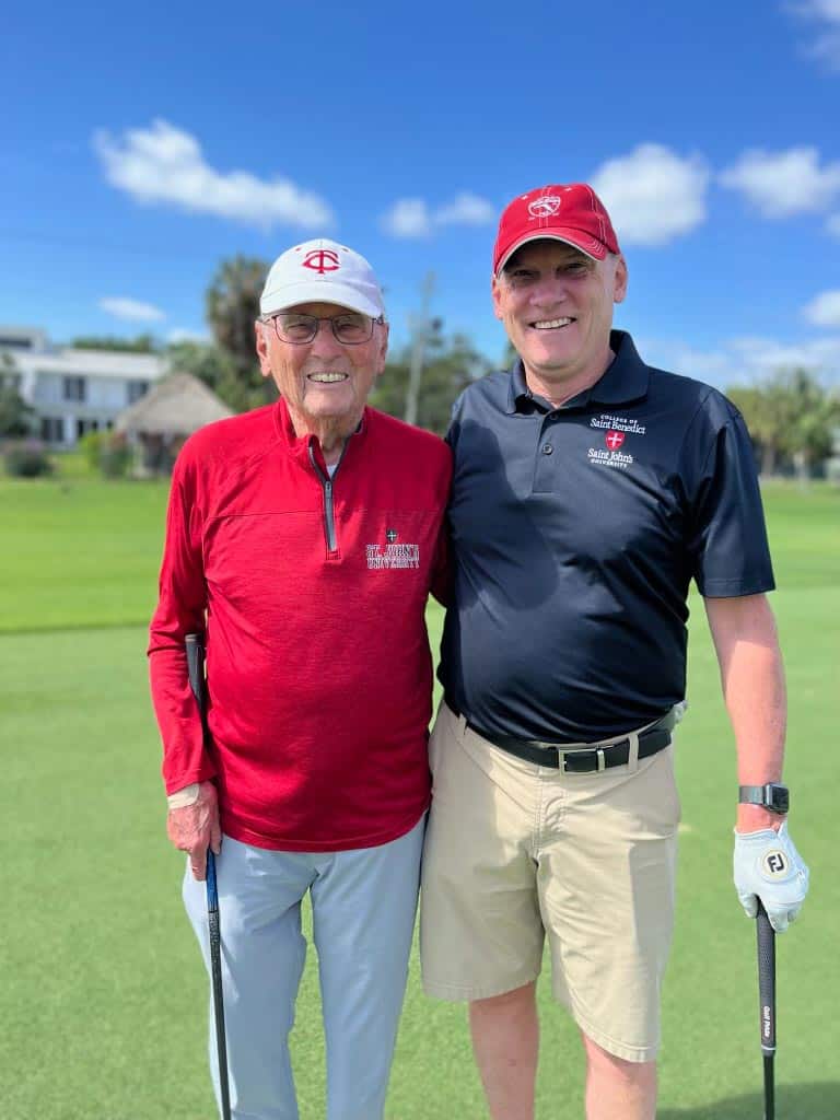 Two people smiling on a golf course. The person on the left is wearing a red long-sleeve shirt, white baseball cap, and light-colored pants, holding a golf club. The person on the right is wearing a black polo shirt, red baseball cap, and beige shorts, also holding a golf club.