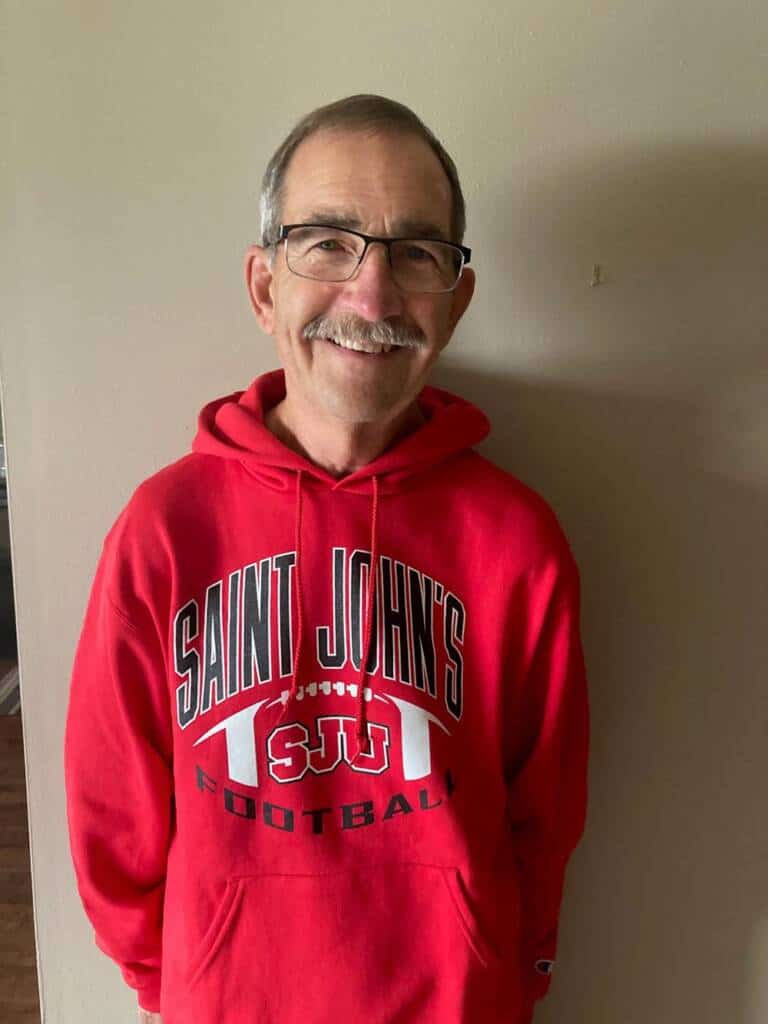 A person with short gray hair and glasses is smiling and standing indoors against a light-colored wall. They are wearing a red hoodie that reads "SAINT JOHN'S FOOTBALL" with the letters "SJU" and a football graphic.