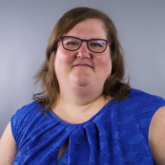 A person with shoulder-length brown hair and glasses is smiling at the camera. They are wearing a blue, textured sleeveless top and are positioned against a plain gray background.