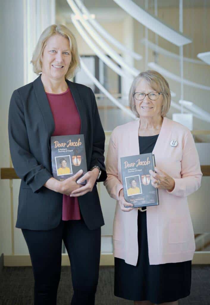 Two women stand side by side, smiling and holding copies of a book titled "Dear Jacob." The woman on the left wears a black blazer over a red top, while the woman on the right wears a light pink cardigan. The background features a modern interior with spiral architectural elements.