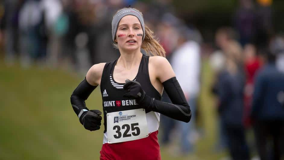 A runner wearing a "Saint Ben" jersey, number 325, is competing in a race. She has black gloves, a gray headband, and race number pinned to her front. Red and black face paint is visible on her cheeks. Blurred spectators are seen in the background.