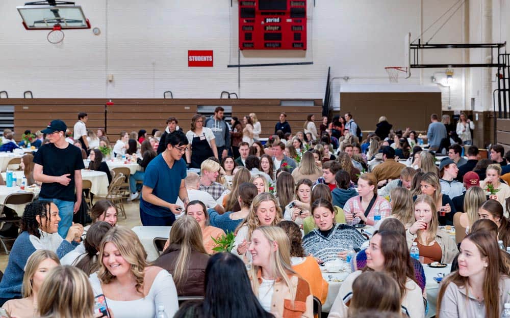 A crowded gymnasium filled with people sitting at tables, many engaged in conversation and some eating. The gym's scoreboard and bleachers are visible in the background. A person wearing a cap is walking among the tables, and decorative plants adorn the scene.