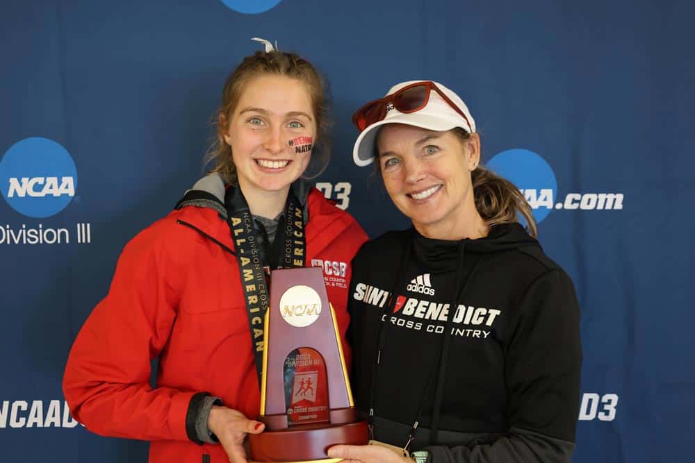 Two women stand together smiling, holding an NCAA Division III trophy. The woman on the left wears a red jacket with a medal around her neck and face paint. The woman on the right wears sunglasses, a white cap, and a black jacket. The backdrop features NCAA logos.