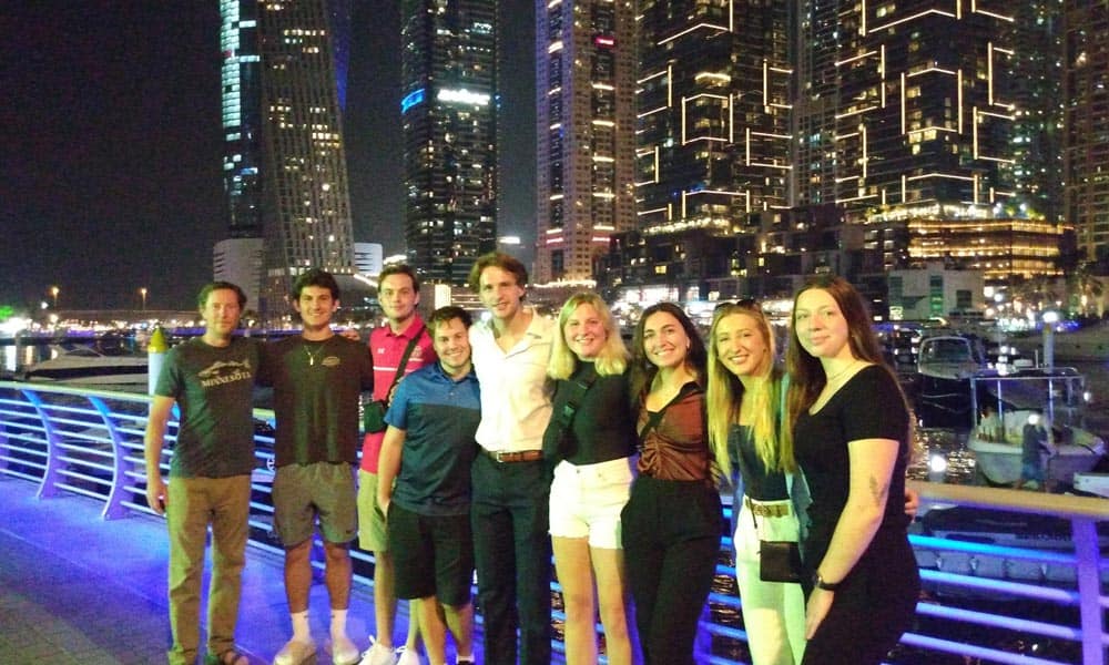 A group of eight people standing side by side and smiling at night in front of an illuminated cityscape with tall, modern buildings. They are on a bridge with railings, and the city lights reflect on the water behind them.