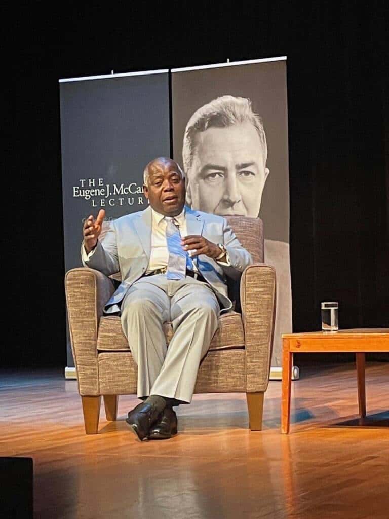 A man in a light gray suit sits on a stage in an armchair, speaking and gesturing with his left hand. Behind him is a black and white banner featuring the Eugene J. McCarthy Lecture and a large image of an older man. A small table with a glass of water is next to him.