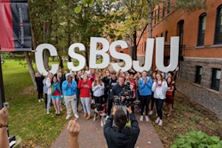 A large group of people stands on a brick path holding up large white letters spelling out "CSBSJU" outside a brick building surrounded by trees. Some individuals have their hands raised in celebration. A person in the foreground is filming the scene.