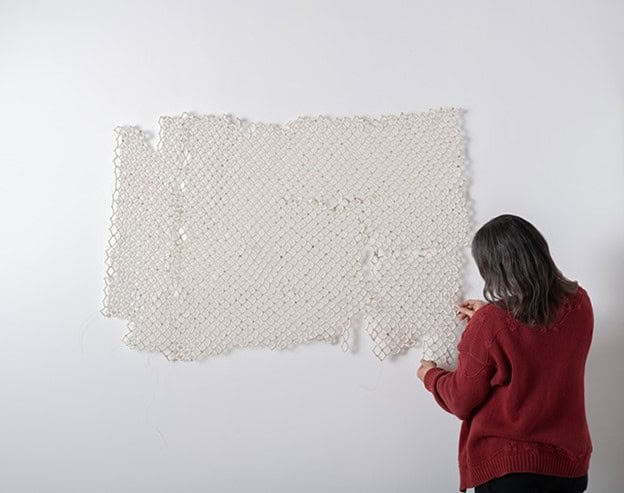 A person with long dark hair, wearing a red sweater, stands against a white wall, working with their hands on a large, textured, rectangular piece of white mesh or netting that is mounted on the wall.