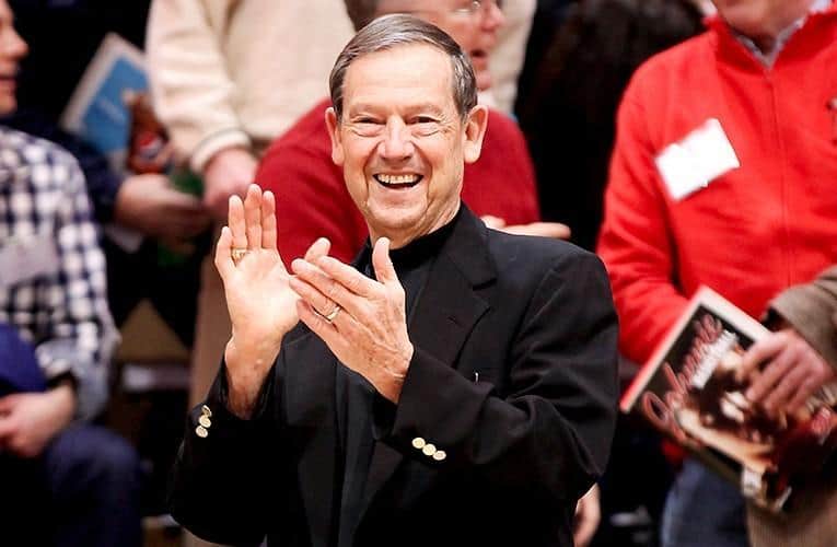 A man in a black suit and glasses smiles and claps his hands. He is in a crowded indoor setting with other people in the background, some holding books or programs.