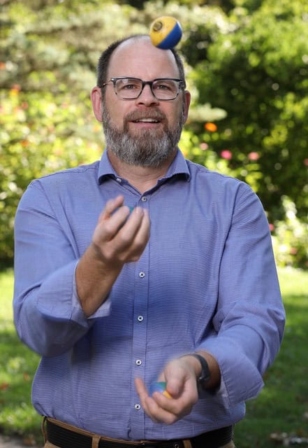 A man with glasses and a beard is standing outdoors in a garden or park, wearing a blue shirt. He is juggling three small balls, one of which is in mid-air. The background is filled with greenery and colorful flowers.