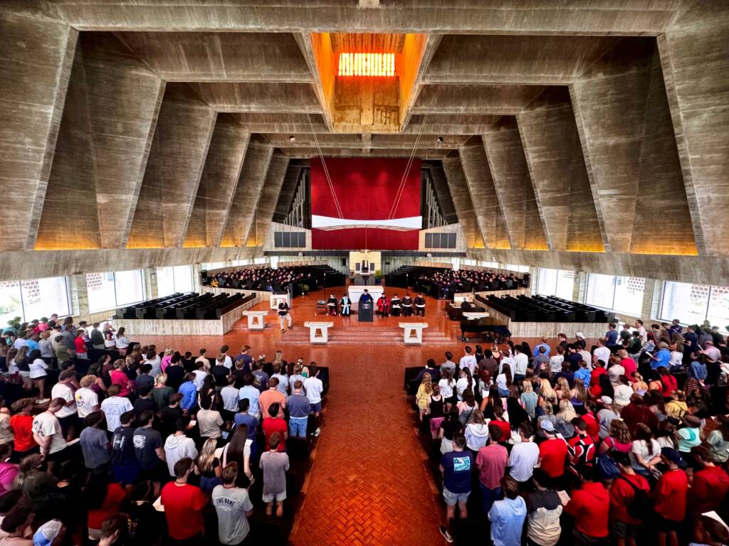 A large gathering of people seated in a spacious modern church with high concrete ceilings. Groups of people are facing the central altar, where clergy members and choir members are positioned. The church is well-lit and has a red backdrop with a white cross.