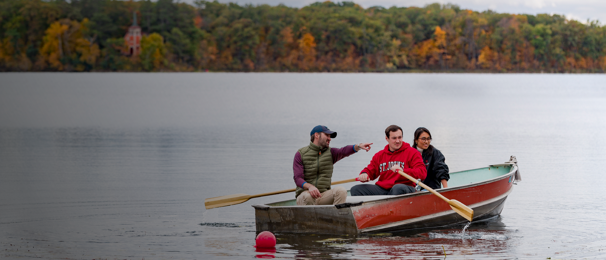 Three people in a small boat on a lake, surrounded by autumn foliage.