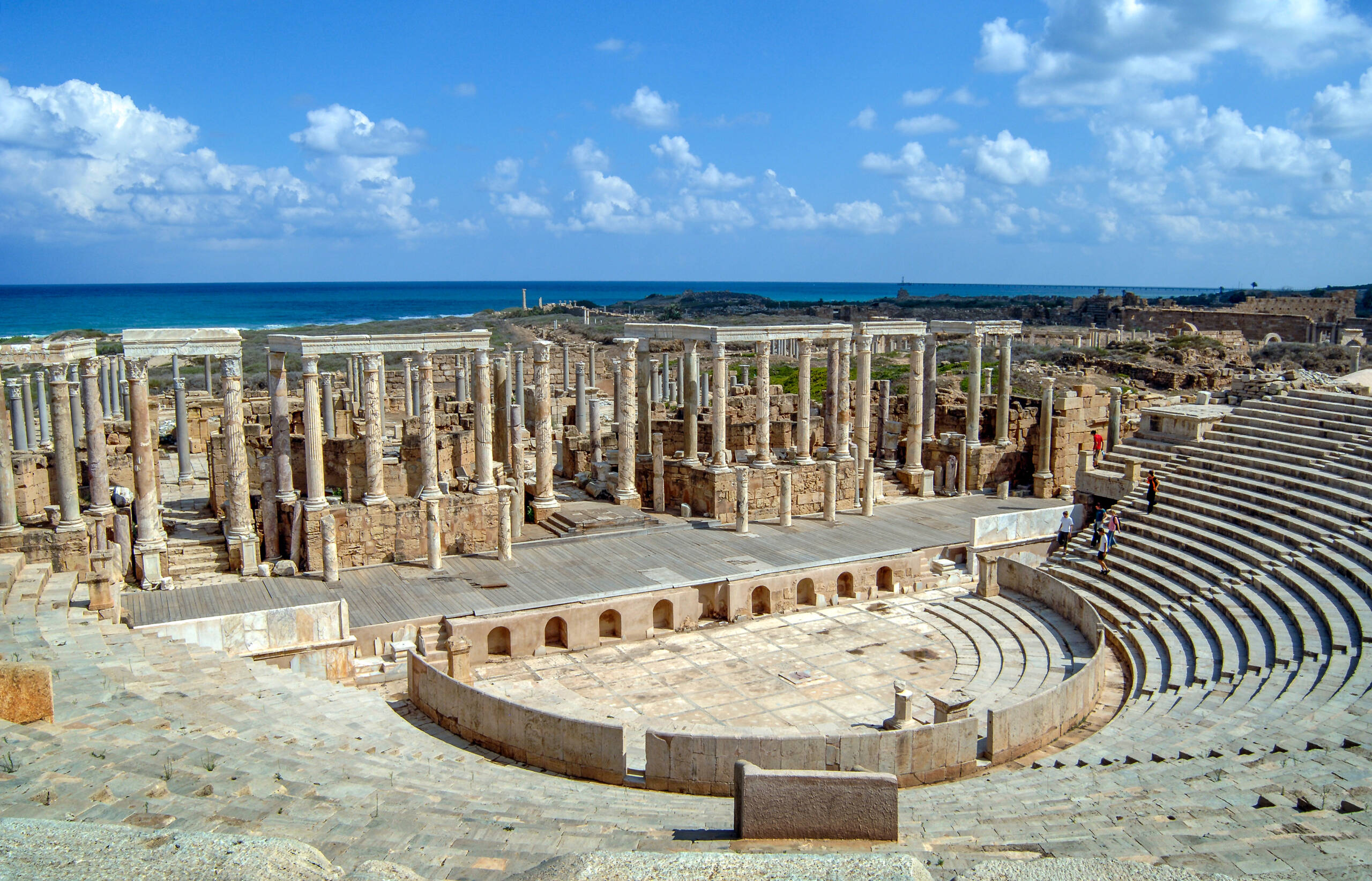 Ancient Roman amphitheater with stone seating and columns, set against a blue sky with scattered clouds. Beyond the ruins, the landscape extends to a distant coastline. The site appears well-preserved, showcasing historical architecture and open space.